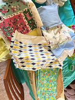 Multiple folded table linens showing pink/green print cloth, blue embroidered cloth, harlequin placemat and teal floral cloth