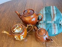 three glazed brown teapots with tea cozy on wooden table