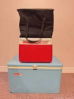 Blue metal Coleman cooler with latch bottom; red plastic Coleman 10 cooler and black soft bag stacked