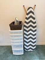 Overall view of iron resting in wicker basket on top of six-drawer plastic tower beside chevron ironing board