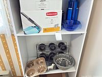 Shelf with Rubbermaid box, blue bowl, tumblers, multiple bake pans, cooling rack, measuring spoons