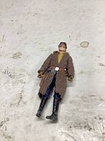 Front view of 3.75-inch figure showing brown trench coat, yellow shirt, blue pants, black boots, belt with holster, goggles on cap