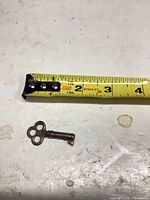 Skeleton key beside tape measure showing length about 1.5 in., visible rust/corrosion