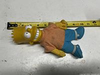 Front view of Bart Simpson plush beside measuring tape showing length