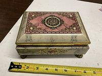 Top view of closed tin showing faux marble, pink panel, floral medallion, tape for scale