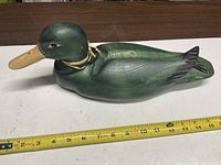 Left side view of painted green wooden duck decoy with tan beak and raffia collar, tape measure shows length