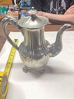 Full view of silver-plated coffee pot showing body, handle, lid, feet, and measuring tape for scale
