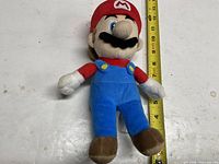 Front view of Mario plush beside tape measure showing size