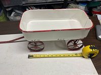 Left side view of white metal wagon with red wheels and handle against measuring tape