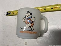 Front view of white milk glass mug with Fred Flintstone and Canada’s Wonderland graphic, tape measure for scale