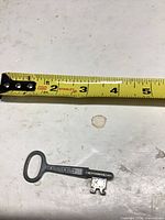 Key beside tape measure showing length about 2.5-3 in., stamped marking visible on shaft