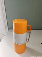 Front view of orange/white Thermos cup, handle visible, THERMOS logo on lid
