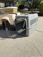Grey plastic pet carrier with door open, partial dog house and bed visible