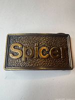 Front of brass belt buckle showing raised "Spicer" lettering and textured background
