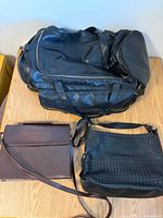Overview of black duffel, brown flap bag, black woven crossbody on table