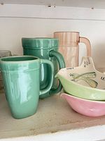 Group view of two seafoam mugs, pink plastic pitcher, two stacked ceramic gravy separators
