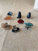 Seven adjustable silver-tone rings displayed on fabric surface, showing variety of stone colours and shapes