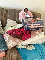 Sofa with patterned fleece throw over stack of assorted throws, boxed UNO Dominoes and Scrabble games on top, crocheted baby doll on backrest