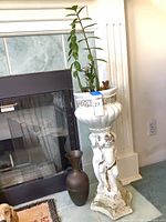 Full view of cherub pedestal planter with plant and metal floor vase beside fireplace