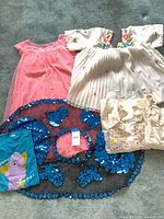Overview of five girls clothing items laid out: pink dress, white embroidered blouse, turquoise graphic tee, blue sequin circular skirt, cream floral skirt.