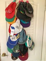 Overall view of approx. 12 adjustable baseball caps hanging on door rack showing colors, logos, and patterns