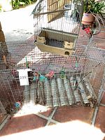 Overall view of both cages; large rectangular cage on bottom, small domed cage placed on top; shows feeders, toys, rust and bent wires