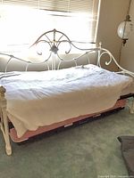 Overall view of white metal daybed showing scrollwork, porcelain finials, mattress and trundle beneath