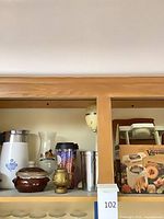 Shelf view showing Corning Ware pieces, Hard Rock glass, stainless mixer cup, brown crock, Veg-o-matic box