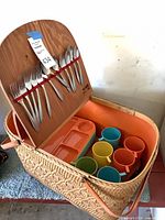 Open wicker basket showing flatware stored in lid, trays and coloured mugs inside