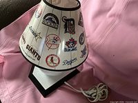 Front view of baseball lamp showing shade with various MLB team logos and baseball-shaped base with cord