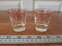 Two Dominion Glass shot glasses with red horizontal measurement stripes beside ruler for scale