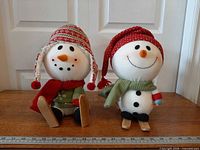 Front view of both plush snowmen showing clothing, faces, and wooden feet/skis