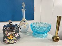 Group shot of blue pressed glass bowl, frosted painted decanter with stopper, enamel biscuit jar with lid, and brass bud vase