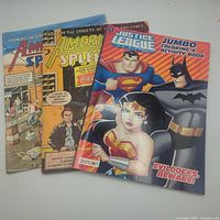 Three publications laid out: two American Splendor comics and one Justice League coloring book
