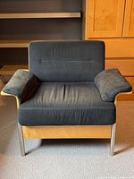 Front view of grey upholstered lounge chair showing seat, backrest, arm pillows, wooden arms and metal legs