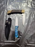 Both folding pocket knives, detached blade and sheath laid out on surface