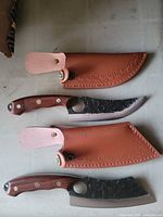 Two knives with hammered carbon steel blades and wood handles accompanied by leather sheaths
