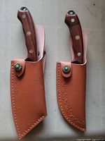 Two knives inserted in leather sheaths, showing wood handles and tooled sheath fronts