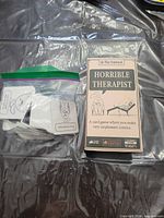 Front of Horrible Therapist NSFW Edition box beside loose cards in zipper bag