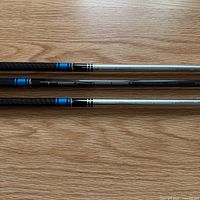 Three shafts laid side by side showing upper portions with blue rings and carbon/silver finishes