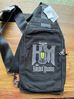 Front view of black Haunted Mansion sling bag showing embroidered logo, strap and hardware