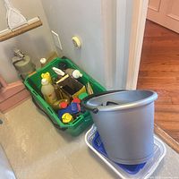 Bucket, dishpan and green tote filled with cleaning products next to dustpan and small appliance