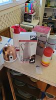 Countertop view showing boxed Carafe Termo, red/white carafe, copper-covered teapot, and Aladdin thermos