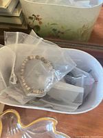 White leatherette case containing plastic bags with a stretch metal bead bracelet; edge of clear glass bird dish visible