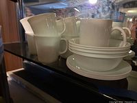Stacked white Corelle mugs, creamer, partial view of plates and bowls