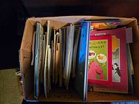 Cardboard box filled with assorted children’s books; visible workbook "What Time Is It?"