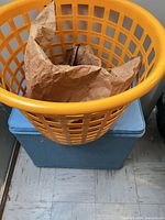 Orange laundry basket sitting on light blue ottoman, paper bag visible inside