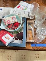 Lot overview showing Pyrex dish, Oneida box, Kitchen Basics package, linens, glasses and ornaments