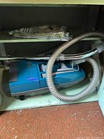 Teal blue and gray canister vacuum with attached hose, metal wand and pack of vacuum bags on shelf
