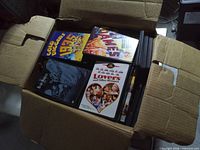 Open cardboard box filled with assorted DVD cases, visible titles Lovers and Other Strangers, Dummies, Code 33 plus additional unidentified cases